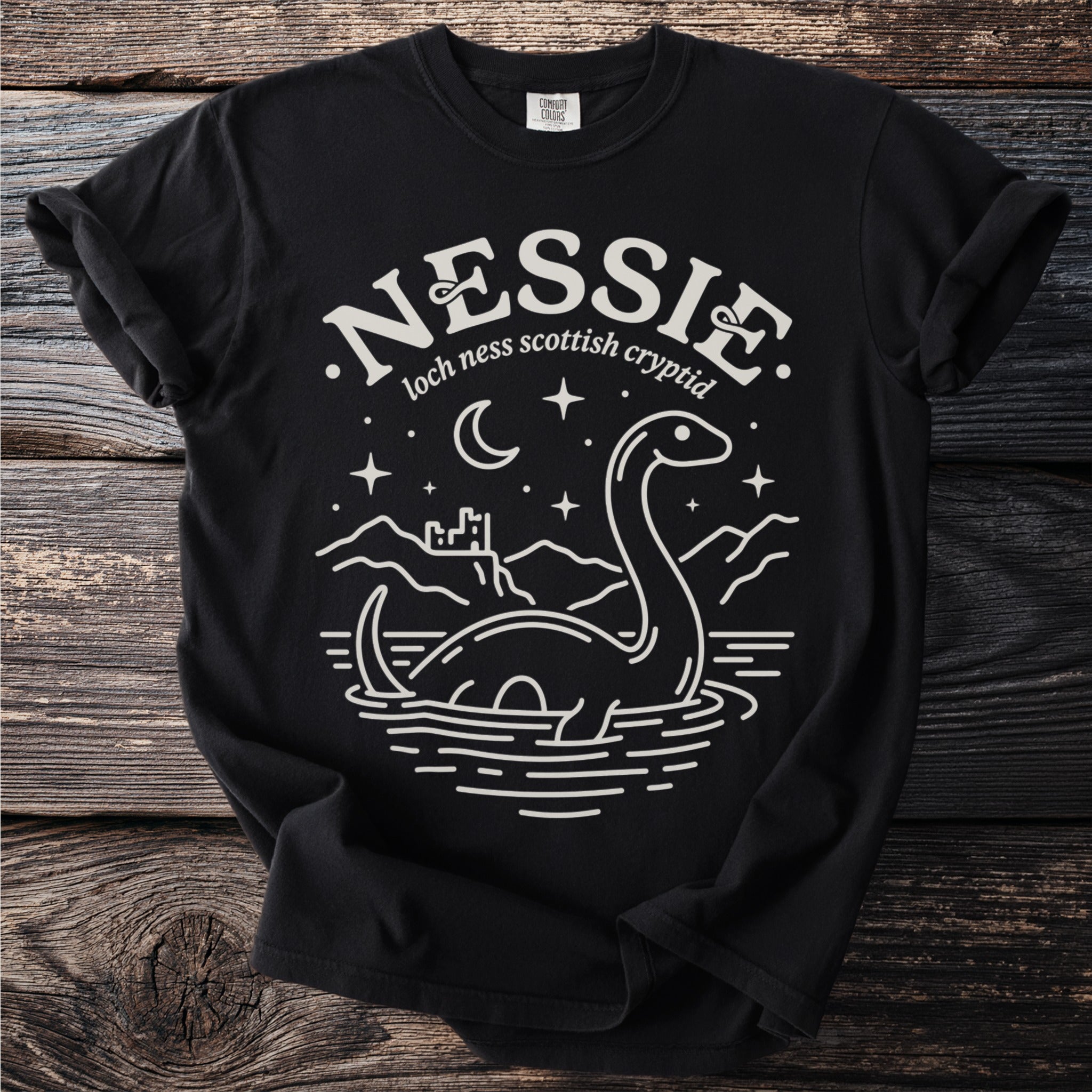 Black t-shirt with a white linework illustration of Nessie, a castle, and mountains.