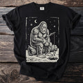 Black t-shirt with a cream folk-cut print: Bigfoot sits on a log with a stick rod over a hole in the ice, one small fish on the line.