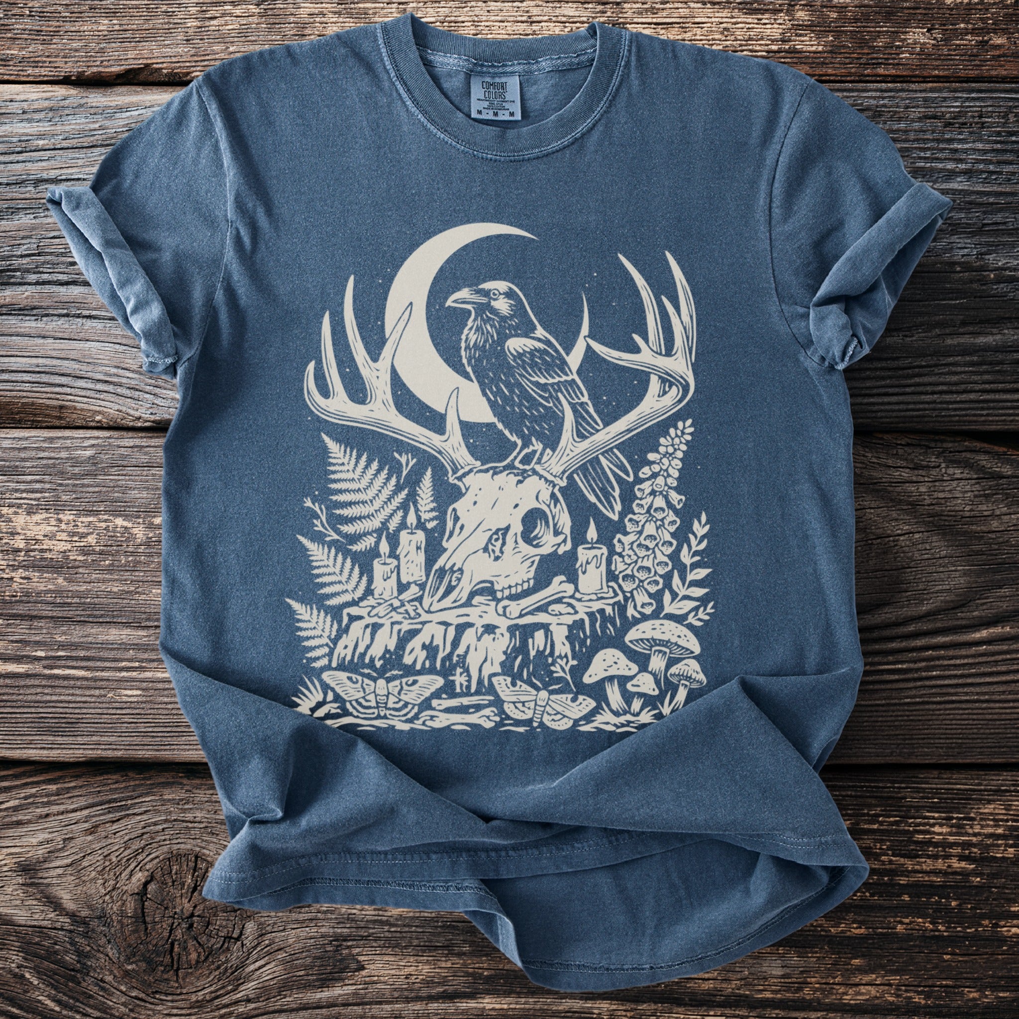 Blue t-shirt with a print of a crow on antlers over a skull, moon, and forest elements.
