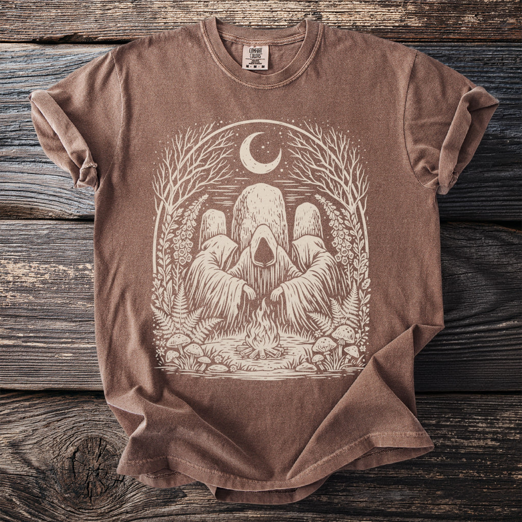 Brown Comfort Colors t-shirt with hooded figures around a campfire under a moon and trees.