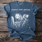 Blue t-shirt with white print of two opossums on a wooden fence and "support local goblins".