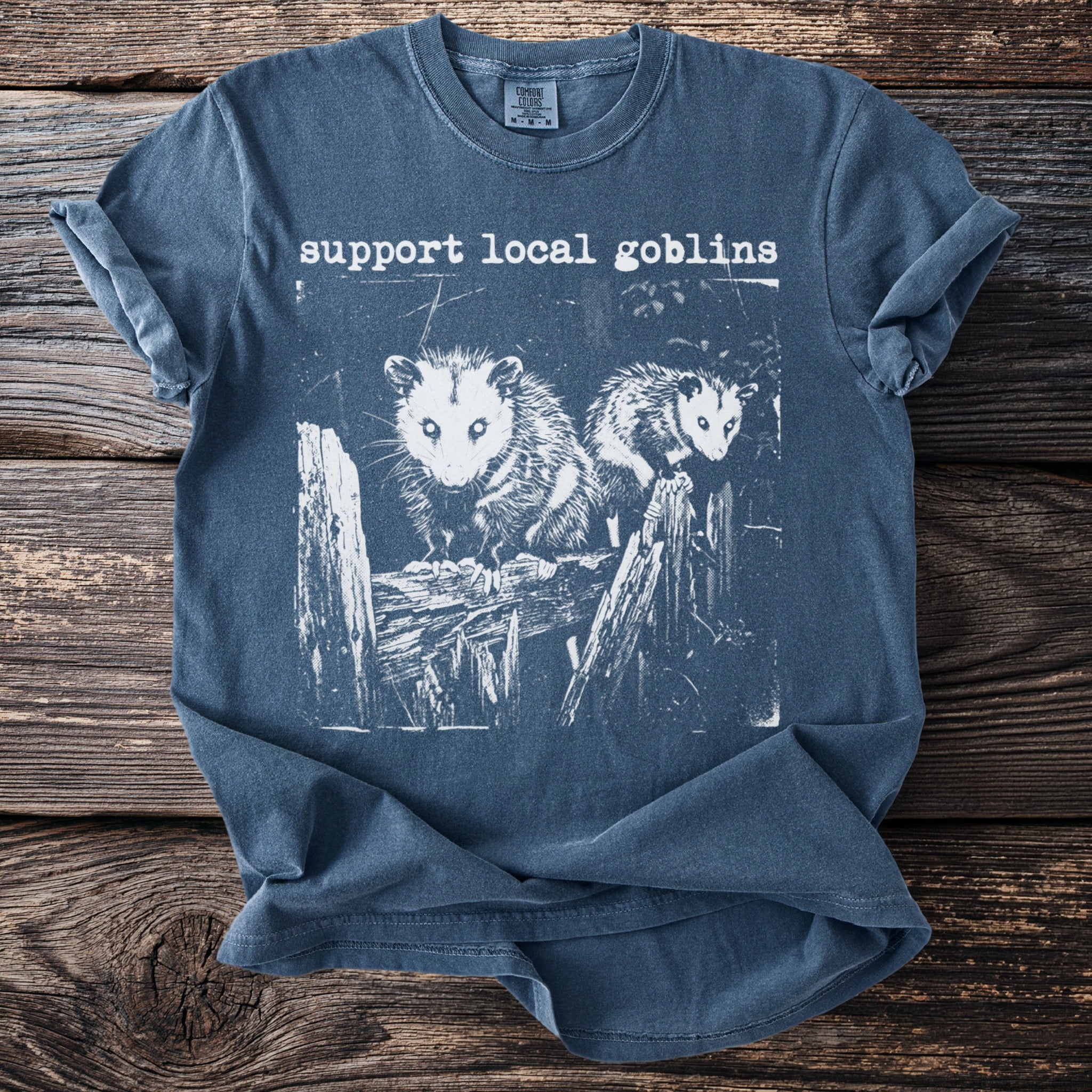 Blue t-shirt with white print of two opossums on a wooden fence and "support local goblins".