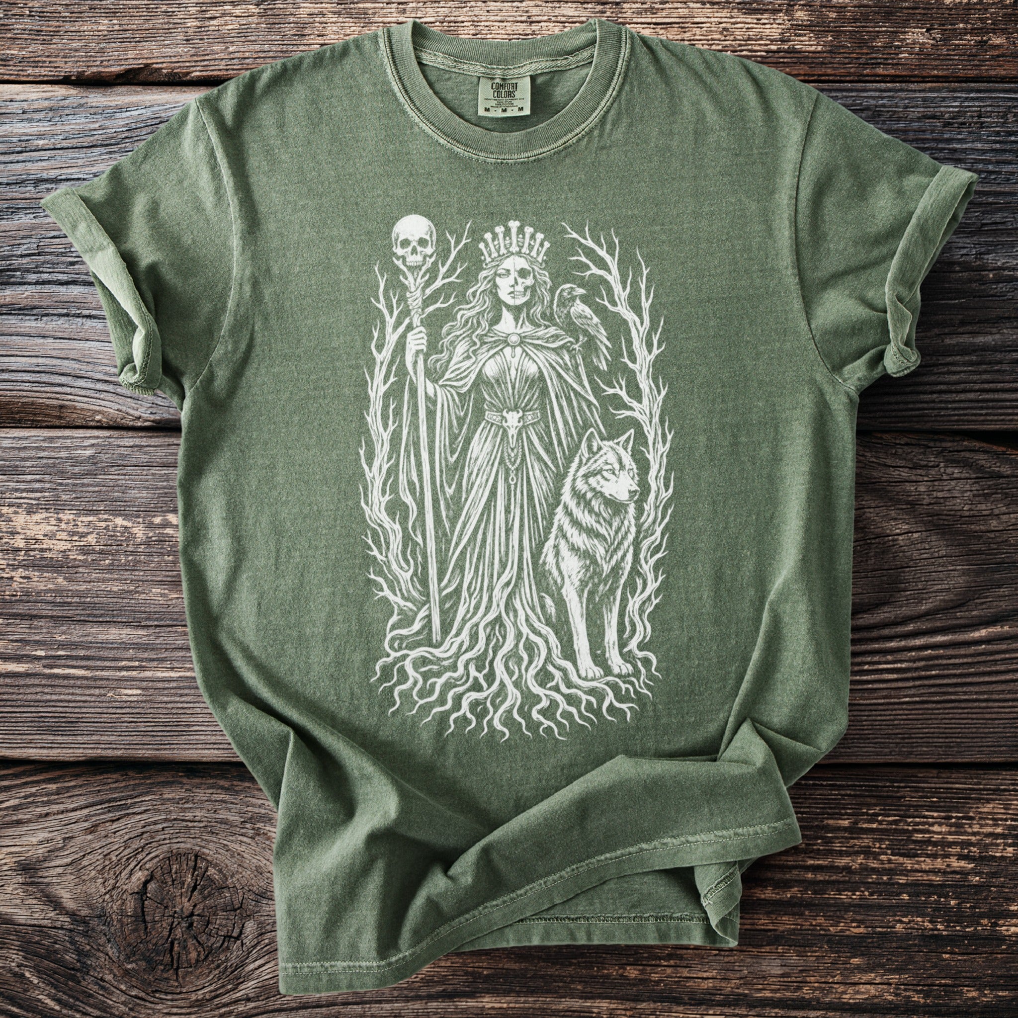 Green t-shirt with white line art of crowned figure, skull staff, wolf, crow, and roots.