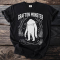 Black t-shirt with white graphic of the Grafton Monster in a wooded, watery landscape.